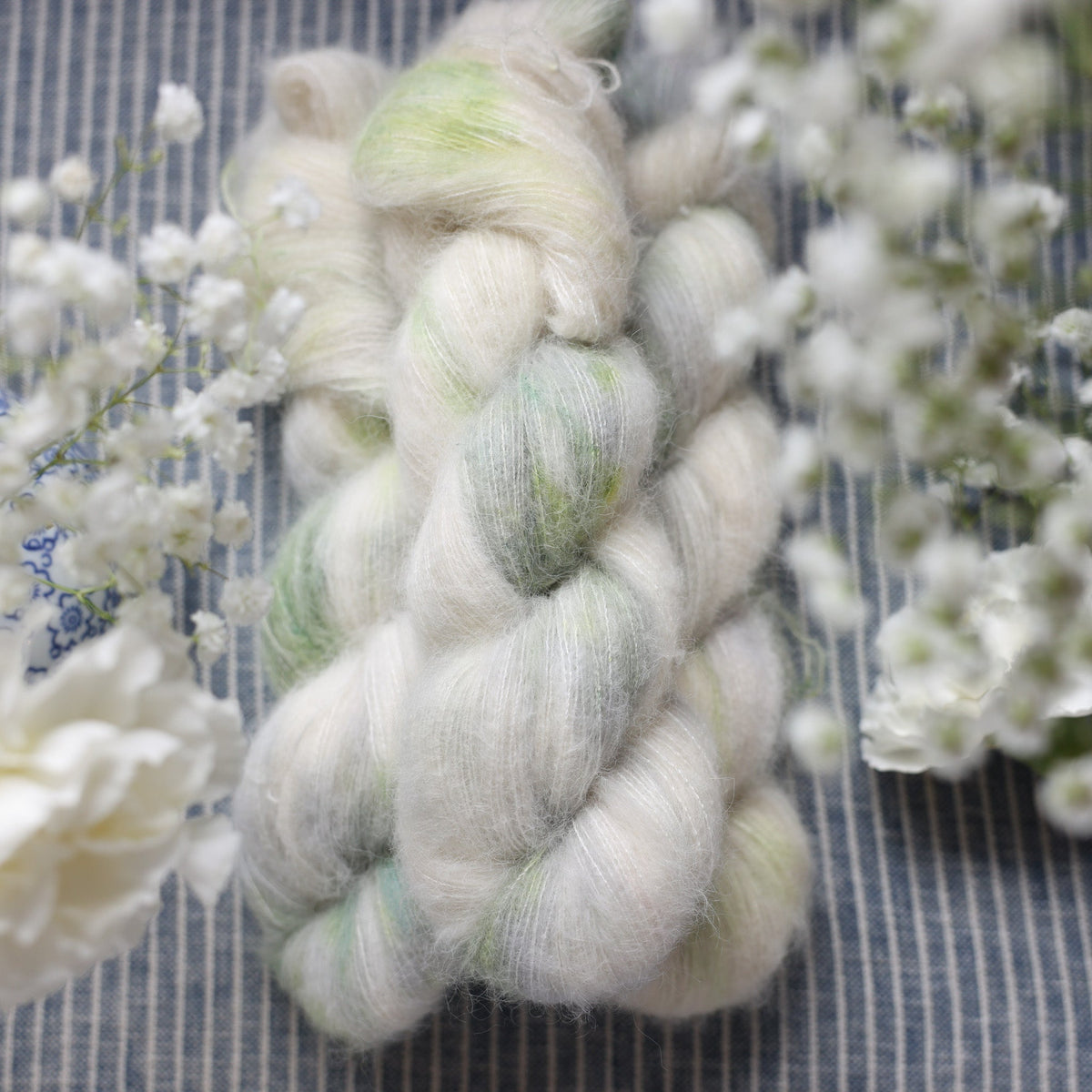 Suri yarn colored a white and very light green with some darker green speckles. These skeins are on top of a faded navy blue and white striped fabric with white flowers like roses and babies breath around the yarn.