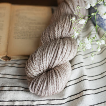A Bulky skein of yarn colored a mist grey tonal-solid. Sitting next to a book and to the right of if there is a blue and white flower pot with white flowers coming out of it. All are sitting on top of a black and with striped table cloth.