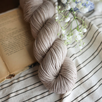 A DK skein of yarn colored a mist grey tonal-solid. Sitting next to a book and to the right of if there is a blue and white flower pot with white flowers coming out of it. All are sitting on top of a black and with striped table cloth.