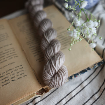 A DK Mini skein of yarn colored a mist grey tonal-solid. Sitting on top of a book book and to the right of if there is a blue and white flower pot with white flowers coming out of it. All are sitting on top of a black and with striped table cloth.
