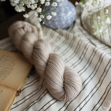 A Fingering skein of yarn colored a mist grey tonal-solid. Sitting next to a book and to the right of if there is a blue and white flower pot with white flowers coming out of it. All are sitting on top of a black and with striped table cloth.