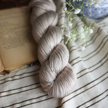 A silk skein of yarn colored a mist grey tonal-solid. Sitting next to a book and to the right of if there is a blue and white flower pot with white flowers coming out of it. All are sitting on top of a black and with striped table cloth.