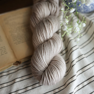 A Sparkle DK skein of yarn colored a mist grey tonal-solid. Sitting next to a book and to the right of if there is a blue and white flower pot with white flowers coming out of it. All are sitting on top of a black and with striped table cloth.