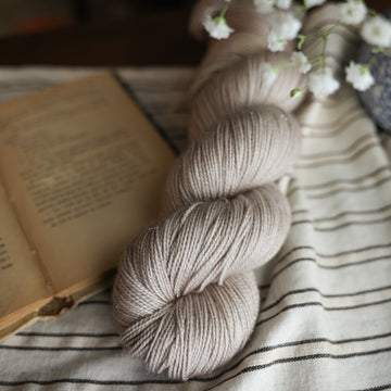A Sparkle fingering skein of yarn colored a mist grey tonal-solid. Sitting next to a book and to the right of if there is a blue and white flower pot with white flowers coming out of it. All are sitting on top of a black and with striped table cloth.