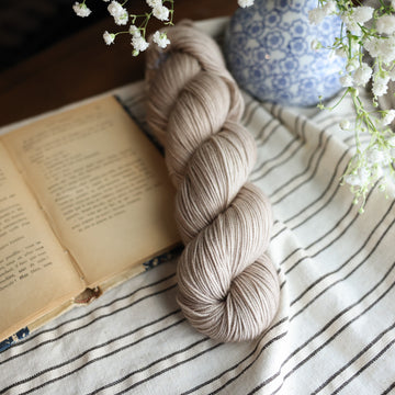 A sport skein of yarn colored a mist grey tonal-solid. Sitting next to a book and to the right of if there is a blue and white flower pot with white flowers coming out of it. All are sitting on top of a black and with striped table cloth.