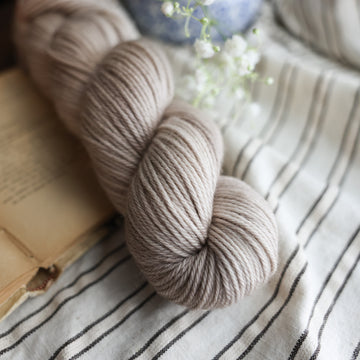 A Worsted skein of yarn colored a mist grey tonal-solid. Sitting next to a book and to the right of if there is a blue and white flower pot with white flowers coming out of it. All are sitting on top of a black and with striped table cloth.