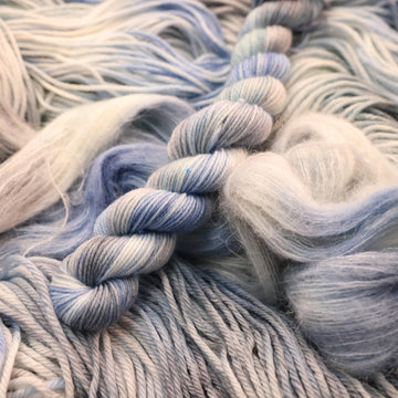 Fingering mini yarn colored white that has very light blue and violet colors mixed in, on top of that same unwound yarn in differed weights.