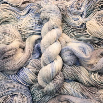 Fingering yarn colored white that has very light blue and violet colors mixed in, on top of that same unwound yarn in differed weights.