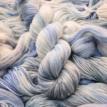 Sport yarn colored white that has very light blue and violet colors mixed in, on top of that same unwound yarn in differed weights.