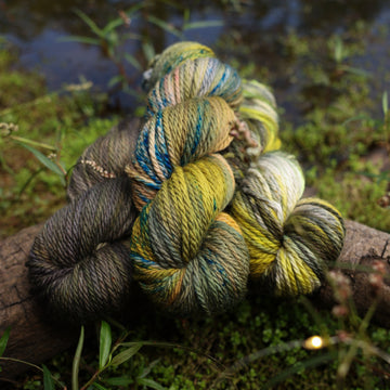 Different Bulky yarn skeins, one colored a lime green and grey, one colored a dark grey, and one colored green with blue speckles, on top of a tree limb that is hovering over a pond, with different mossy plants around the pond, and around the yarn.