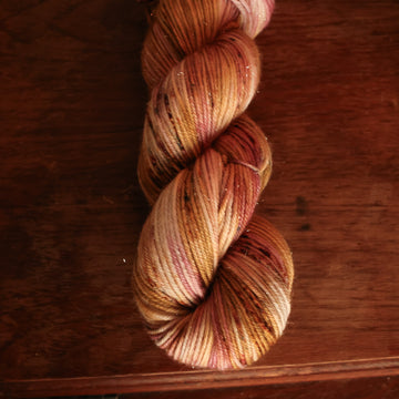 DK sparkle yarn with shades of yellow and pink with dark brown and blue speckles, on top of a wooden table with a plant peeking out of the frame.