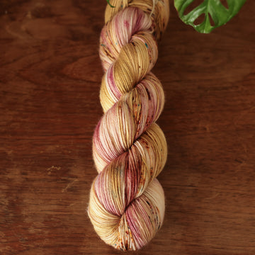 Fingering yarn with shades of yellow and pink with dark brown and blue speckles, on top of a wooden table with a plant peeking out of the frame.
