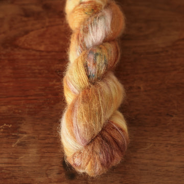 Suri Alpaca yarn with shades of yellow and pink with dark brown and blue speckles, on top of a wooden table with a plant peeking out of the frame.