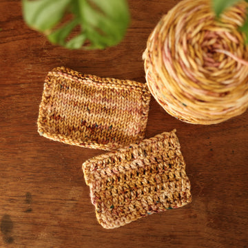 Crochet and Knit swatches with shades of yellow and pink with dark brown and blue speckles, on top of a wooden table with a plant peeking out of the frame, and a caked version of this yarn in the corner.