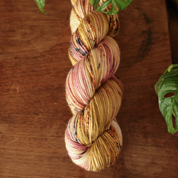 Worsted yarn with shades of yellow and pink with dark brown and blue speckles, on top of a wooden table with a plant peeking out of the frame.