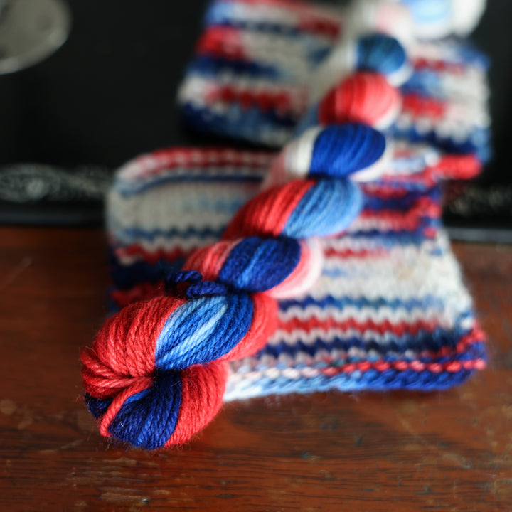 DK mini red white and blue yarn skeins on top crochet and knit swatches of this color way chic is on top of an old fashioned sewing table.
