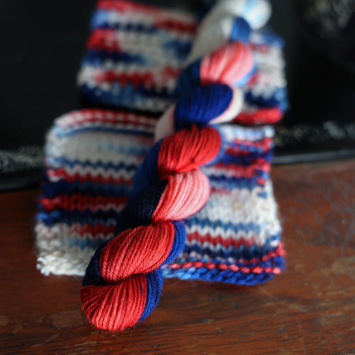 Fingering mini red white and blue yarn skeins on top of knit and crochet swatches also on top of an old fashioned sewing table.