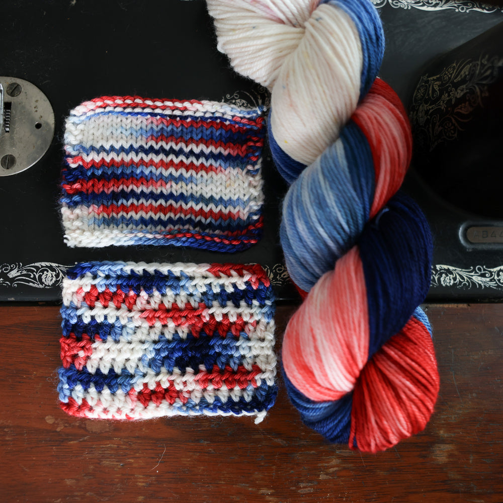Crochet and knit swatches of red white and blue yarn as well as a regular skein of this color way on top of an old fashioned sewing table.