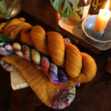 Two skeins of yarn one is a multi colored fingering mini and the second is a golden fingering skein. Both are sitting on top of hand knit socks made of both skeins. All sitting on top of a wooden table.