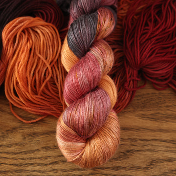 a light sunset orange, red and purple silk yarn with a regular version of it, unwound underneath it, on a wooden table.