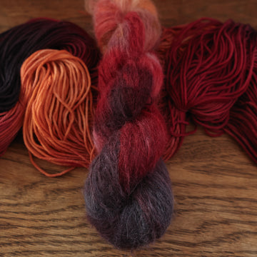 a sunset orange, red and dark purple soft suri yarn, with another regular version of it unwound underneath it, on a wooden table.