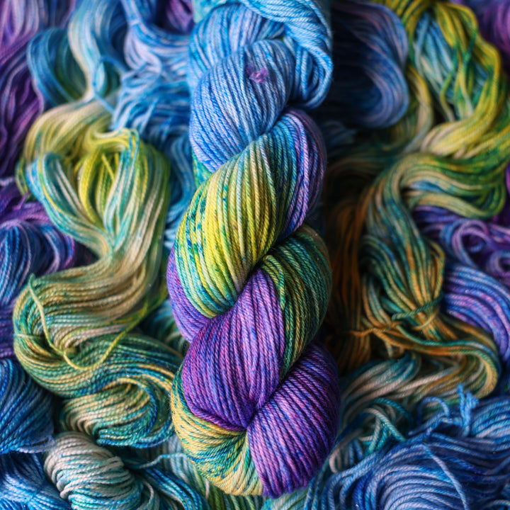 DK yarn colored different shades of light and dark blue along with purple and some yellows and orange and some green. This is on top of unwound skeins of this same color way.