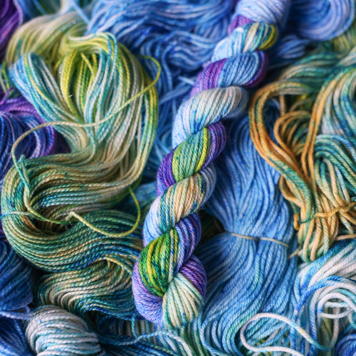 DK mini yarn colored different shades of light and dark blue along with purple and some yellows and orange and some green. This is on top of unwound skeins of this same color way.