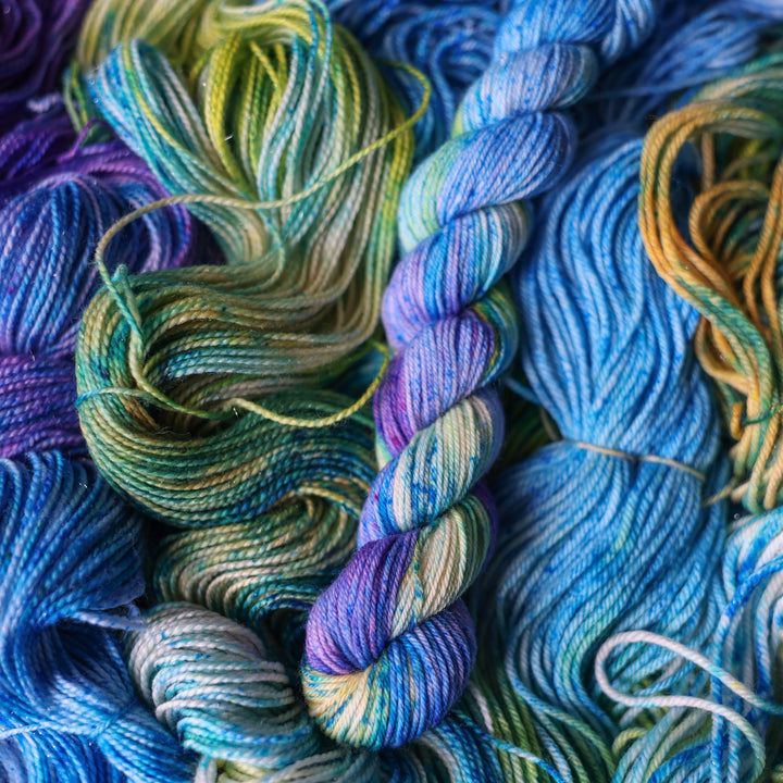 Fingering mini yarn colored different shades of light and dark blue along with purple and some yellows and orange and some green. This is on top of unwound skeins of this same color way.