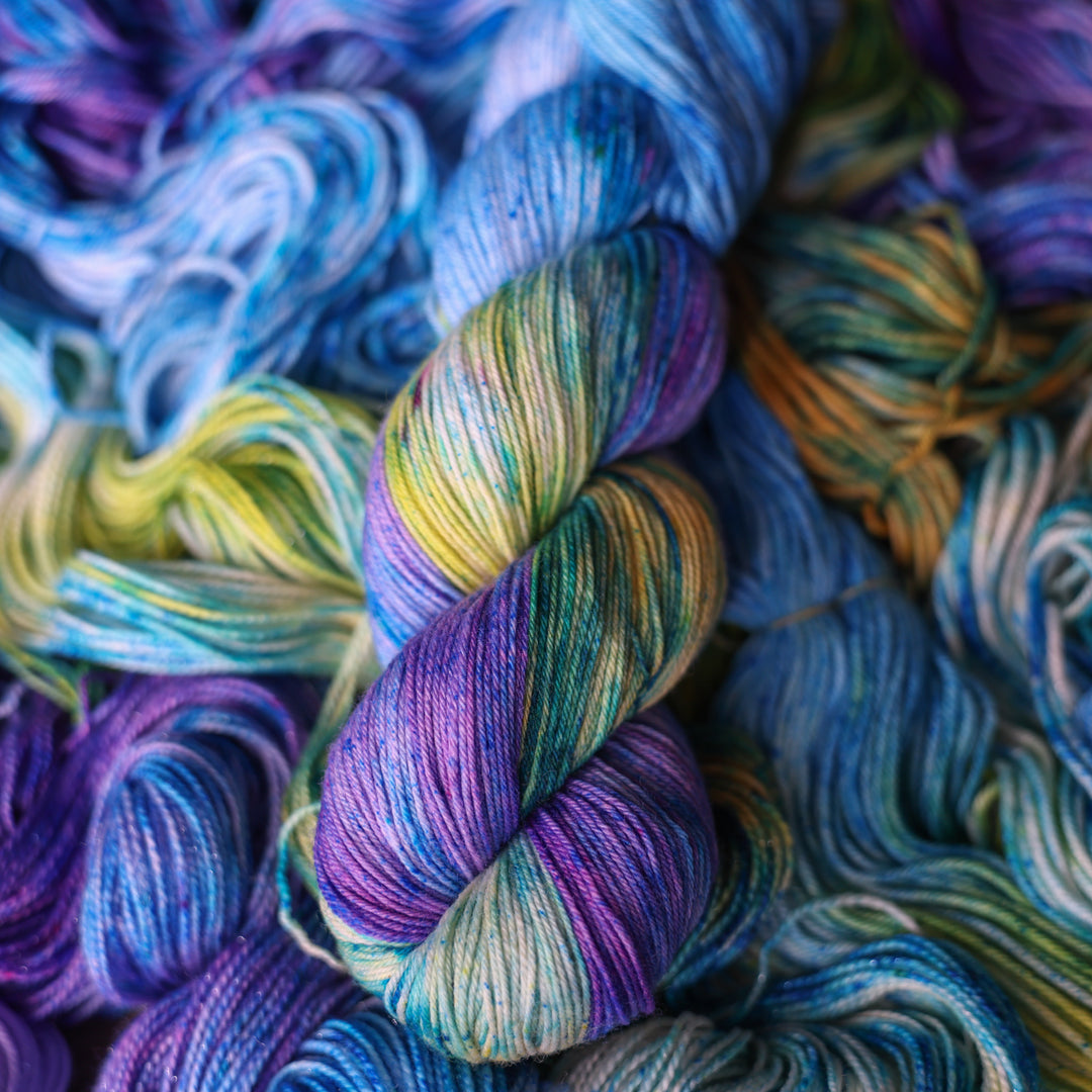 Fingering yarn colored different shades of light and dark blue along with purple and some yellows and orange and some green. This is on top of unwound skeins of this same color way.