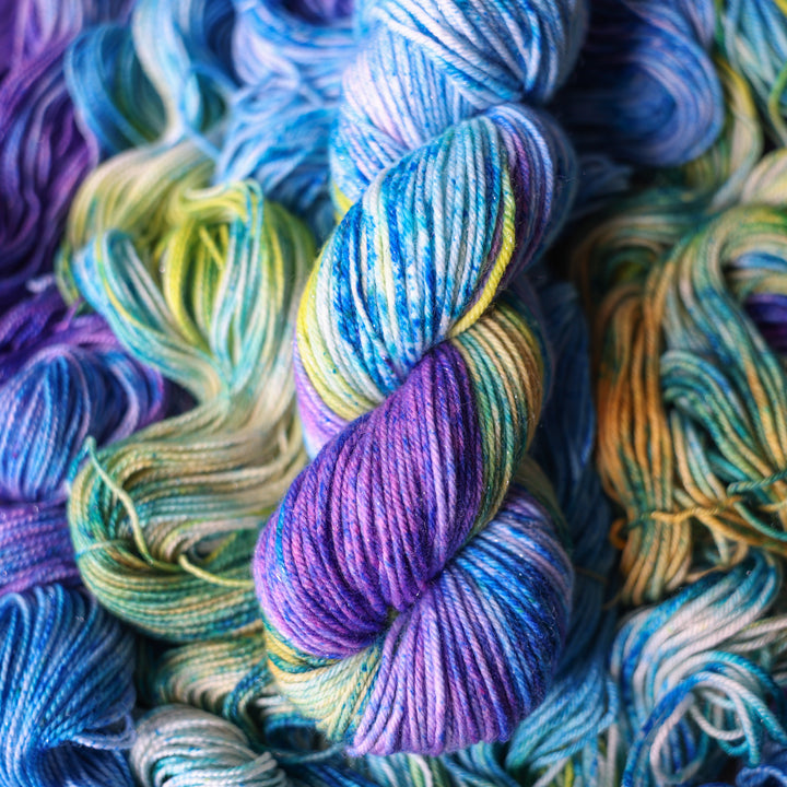Sparkle DK yarn colored different shades of light and dark blue along with purple and some yellows and orange and some green. This is on top of unwound skeins of this same color way.