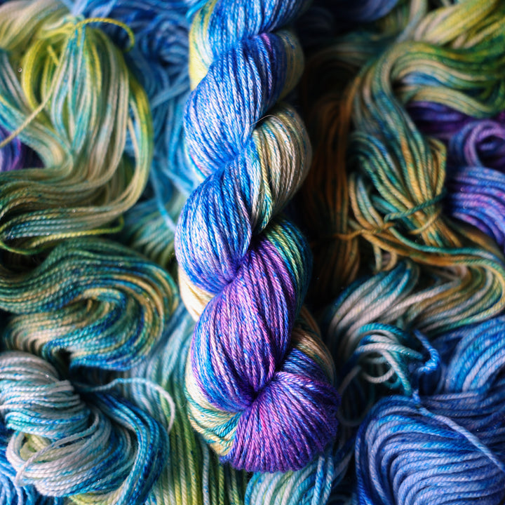 Silk DK yarn colored different shades of light and dark blue along with purple and some yellows and orange and some green. This is on top of unwound skeins of this same color way.