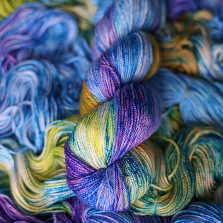 Sport yarn colored different shades of light and dark blue along with purple and some yellows and orange and some green. This is on top of unwound skeins of this same color way.