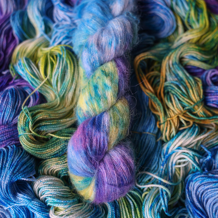 Suri yarn colored different shades of light and dark blue along with purple and some yellows and orange and some green. This is on top of unwound skeins of this same color way.