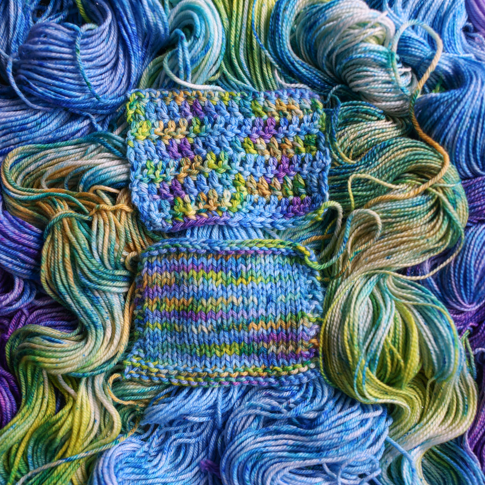 crochet and knit swatches colored different shades of light and dark blue along with purple and some yellows and orange and some green. This is on top of unwound skeins of this same color way.