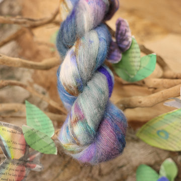 A soft alpaca yarn with a purple, ocean blue and dark green with other similar shades of colors and some speckles on it, on top of a plant with bare branches and leaves in the back. along with painted butterflies and leaves made of book pages, all on top of a wooden table.