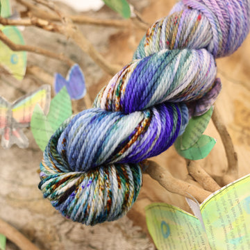 Bulky yarn with a purple, ocean blue and dark green with other similar shades of colors and some speckles on it, on top of a plant with bare branches and leaves in the back. along with painted butterflies and leaves made of book pages, all on top of a wooden table.