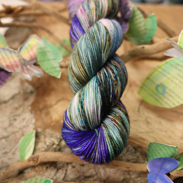 DK yarn with a purple, ocean blue and dark green with other similar shades of colors and some speckles on it, on top of a plant with bare branches and leaves in the back. along with painted butterflies and leaves made of book pages, all on top of a wooden table.