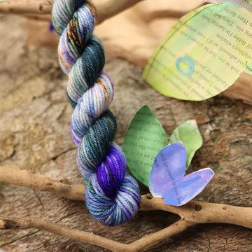 DK mini yarn with a purple, ocean blue and dark green with other similar shades of colors and some speckles on it, on top of a plant with bare branches and leaves in the back. along with painted butterflies and leaves made of book pages, all on top of a wooden table.