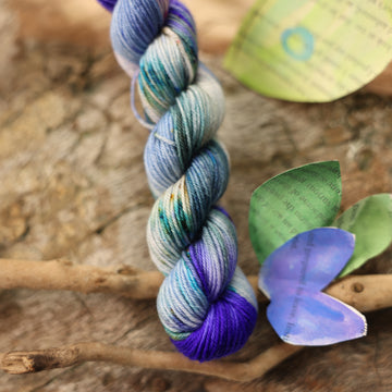 Fingering mini yarn with a purple, ocean blue and dark green with other similar shades of colors and some speckles on it, on top of a plant with bare branches and leaves in the back. along with painted butterflies and leaves made of book pages, all on top of a wooden table.