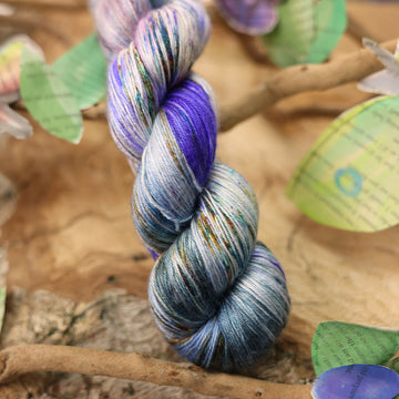Silk yarn with a purple, ocean blue and dark green with other similar shades of colors and some speckles on it, on top of a plant with bare branches and leaves in the back. along with painted butterflies and leaves made of book pages, all on top of a wooden table.