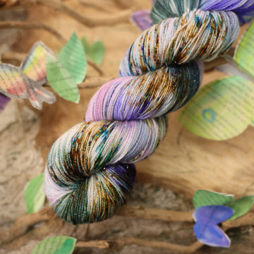 Sparkle yarn with a purple, ocean blue and dark green with other similar shades of colors and some speckles on it, on top of a plant with bare branches and leaves in the back. along with painted butterflies and leaves made of book pages, all on top of a wooden table.