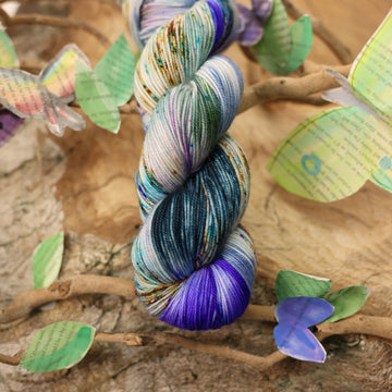 Sport yarn with a purple, ocean blue and dark green with other similar shades of colors and some speckles on it, on top of a plant with bare branches and leaves in the back. along with painted butterflies and leaves made of book pages, all on top of a wooden table.