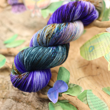 Square fingering yarn with a dark purple, ocean blue and dark green with other similar shades of colors and some speckles on it, on top of a plant with bare branches and leaves in the back. along with painted butterflies and leaves made of book pages, all on top of a wooden table.