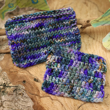 Knit and crochet swatches with a purple, ocean blue and dark green with other similar shades of colors and some speckles on it, on top of a plant with bare branches and leaves in the back. along with painted butterflies and leaves made of book pages, all on top of a wooden table.