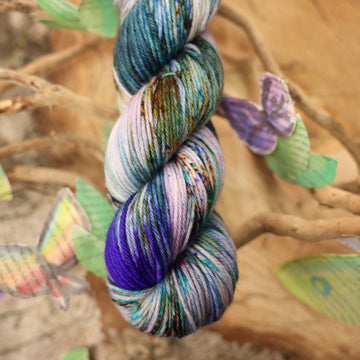 Worsted yarn with a purple, ocean blue and dark green with other similar shades of colors and some speckles on it, on top of a plant with bare branches and leaves in the back. along with painted butterflies and leaves made of book pages, all on top of a wooden table.