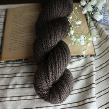 A bulky skein of yarn colored dark, green/brown tonal-solid. Sitting on top a book and black and white striped table cloth. to the side there is a blue and white flower pot with a white flower sticking out over the book. All sitting on a dark wooden table.