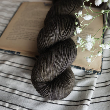 A sport skein of yarn colored dark, green/brown tonal-solid. Sitting on top a book and black and white striped table cloth. to the side there is a blue and white flower pot with a white flower sticking out over the book. All sitting on a dark wooden table.