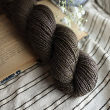 A Worsted skein of yarn colored dark, green/brown tonal-solid. Sitting on top a book and black and white striped table cloth. to the side there is a blue and white flower pot with a white flower sticking out over the book. All sitting on a dark wooden table.