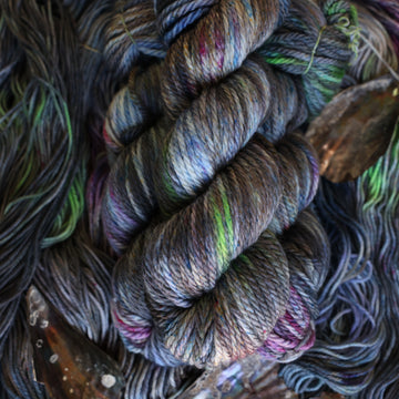 Bulky yarn colored with different shades of grey and streaks of green, and dark pink, also there are also blue and purple speckles. These skeins are on top of unwound skeins of the same color way with some shells on top.