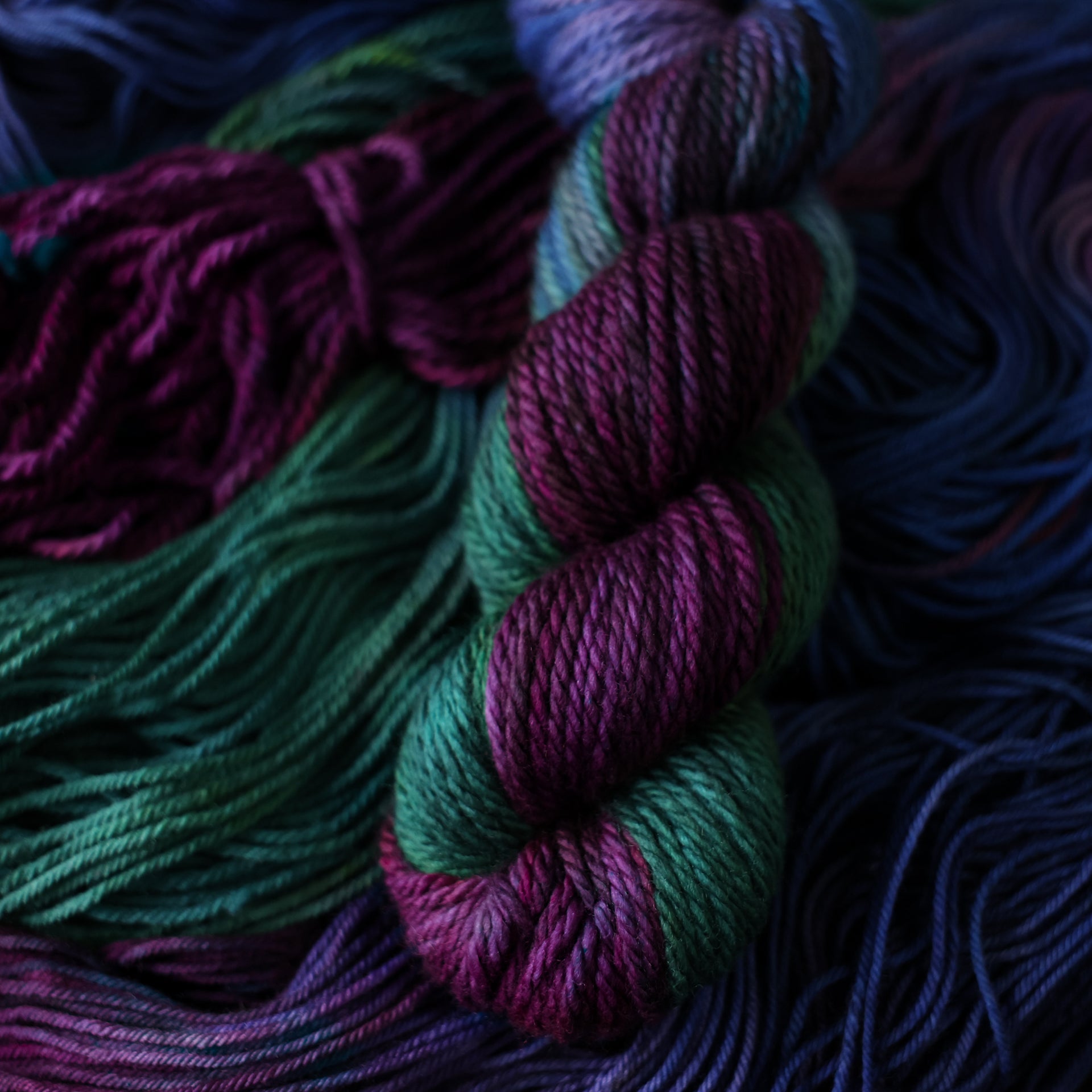 Bulky yarn colored with an emerald green, Dark blue, and dark purple, on top of un unwound version of that yarn in a different weight.
